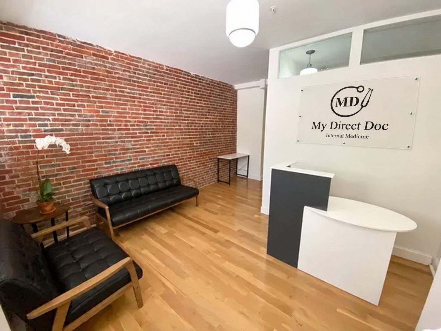 Reception area of My Direct Doc with a brick wall, black leather seating, wood floors and the My Direct Doc sign above the front desk.